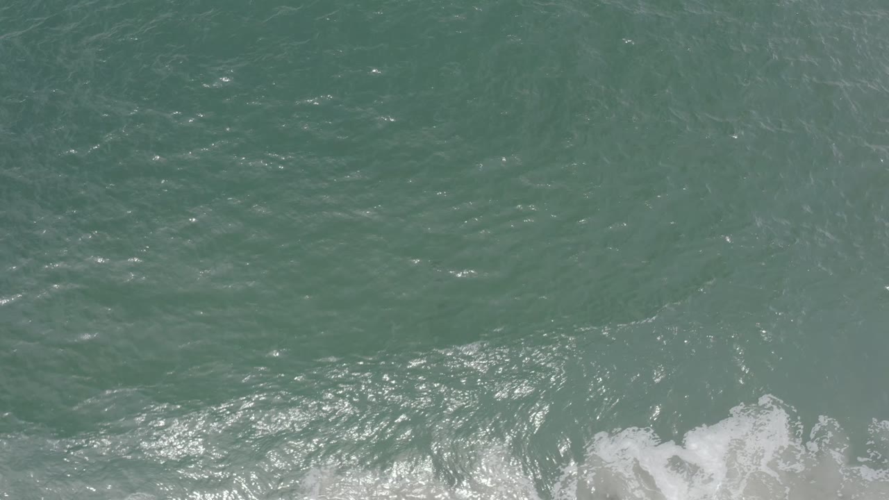 Overhead Los Cabos water surface with green-blue ripples, scattered sunlight highlights, low white foam entering from the bottom edge, and no visible people.