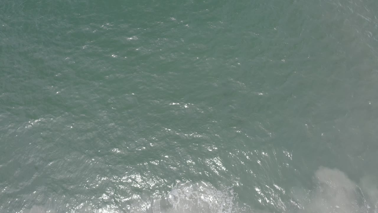 Top-down Los Cabos ocean frame with brighter sun glints across the water and soft pale submerged forms near the lower edge, no visible people.