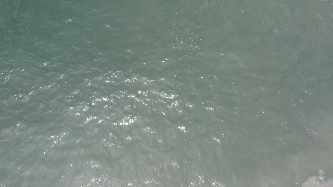 Aerial view of gray green water in Los Cabos, Mexico, where bright ripples cluster across the middle and pale foam remains near the lower edge with no visible people.