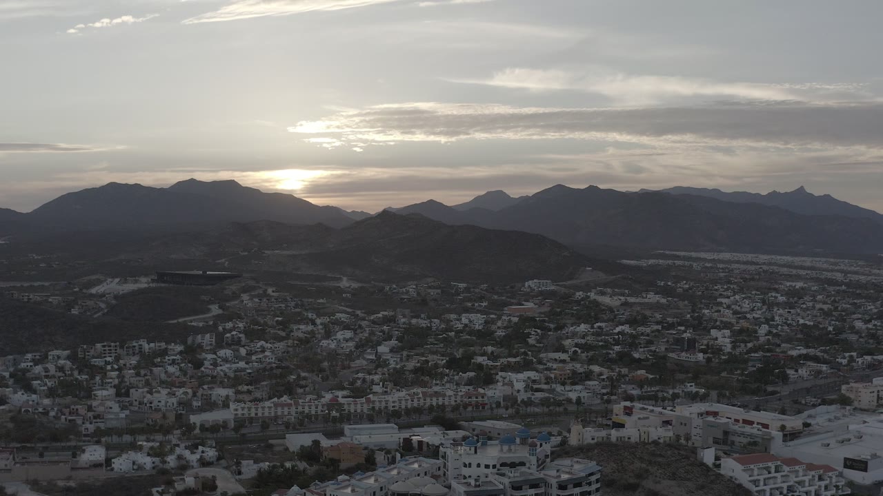 Los Cabos foothill community seen from above with white buildings, dark mountain ridges, gray sunrise clouds, and no visible people.