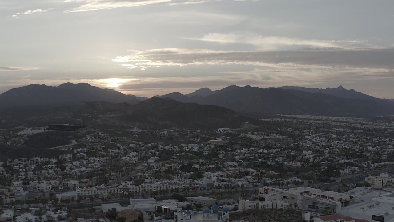 Aerial city view below dark mountains with low sun behind clouds.