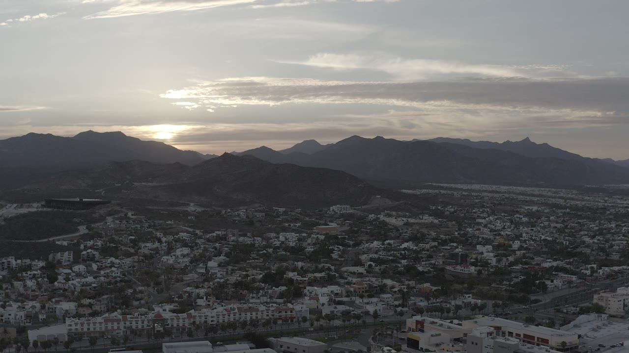 Aerial view over Los Cabos, Mexico, with a town in shadow below dark hills, a mountain range on the horizon, the sun low behind clouds, and no visible people.