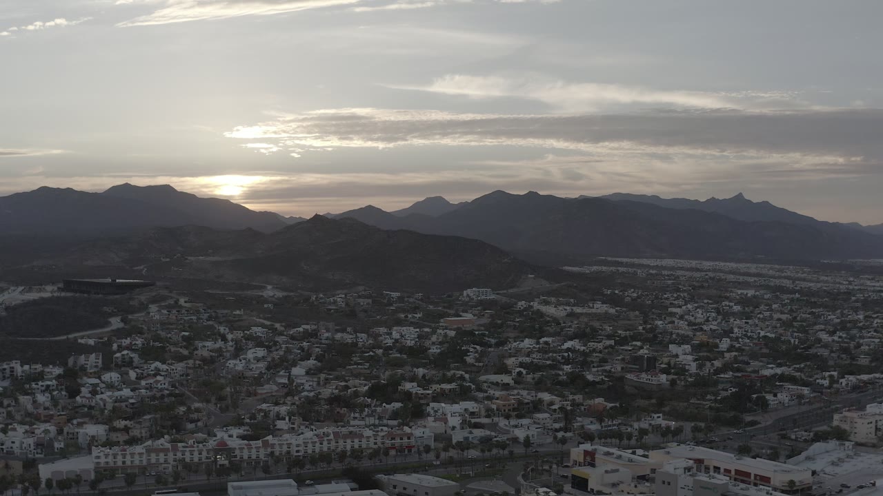 Elevated Los Cabos view with clustered buildings across the lower frame, dark desert hills in the middle distance, layered mountains beyond, and no visible people.
