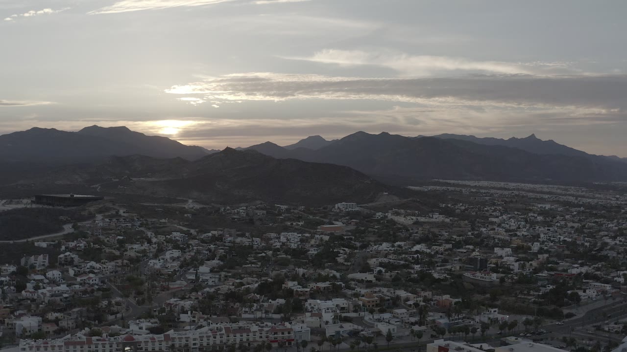 Los Cabos rooftops and roads sit in the lower frame while layered mountains and a muted sunset sky fill the distance, no visible people.