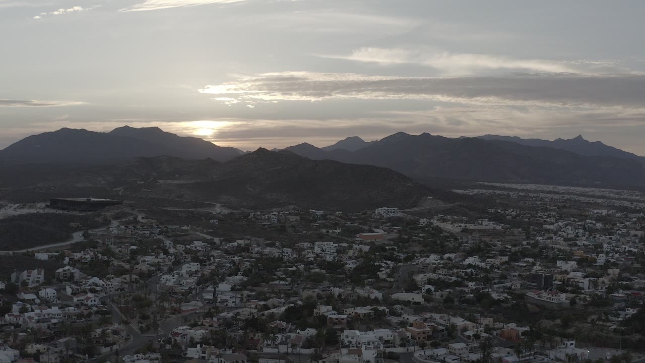 Opening aerial frame over Los Cabos, Mexico, with hillside homes below silhouetted desert ridges, a bright low sun behind clouds, and no visible people.