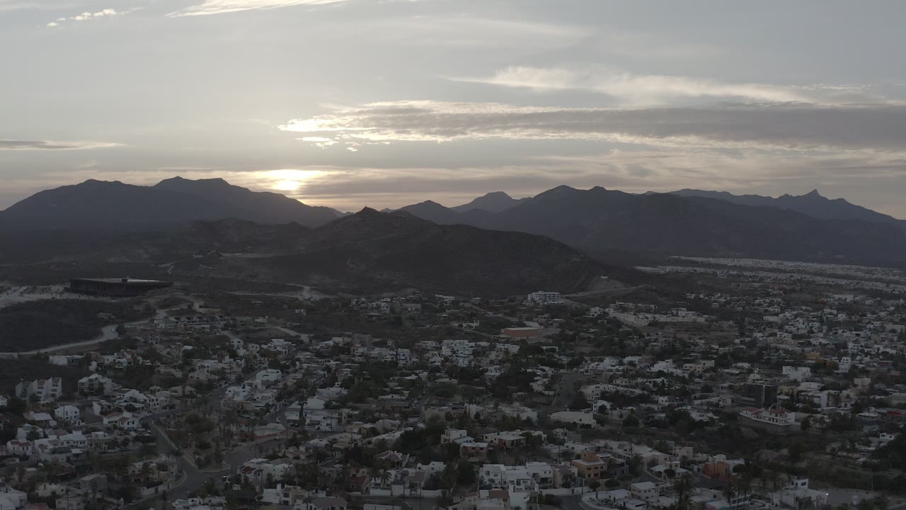 Aerial view in Los Cabos, Mexico, showing more town rooftops and roads beneath a clouded sky, with dark mountain silhouettes and no visible people.
