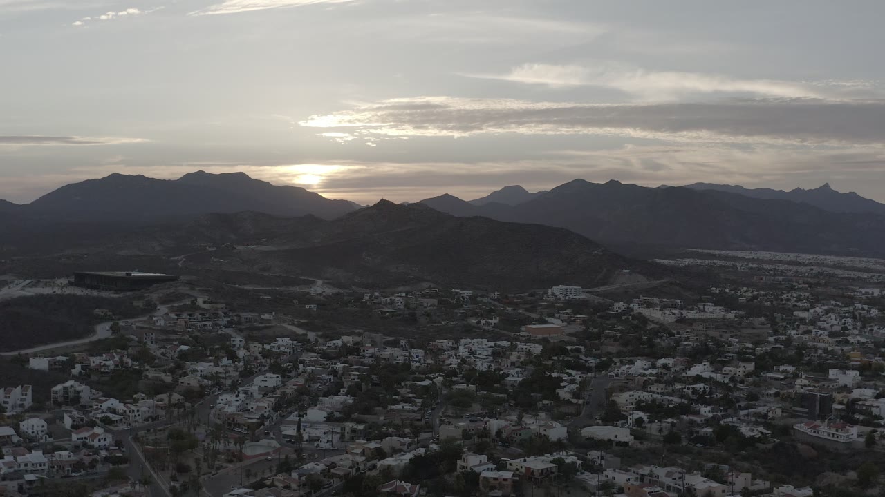 Los Cabos aerial frame with clustered buildings in shadow, low hills across the middle of the frame, distant mountains, gray clouds, warm light near the horizon, and no visible people.