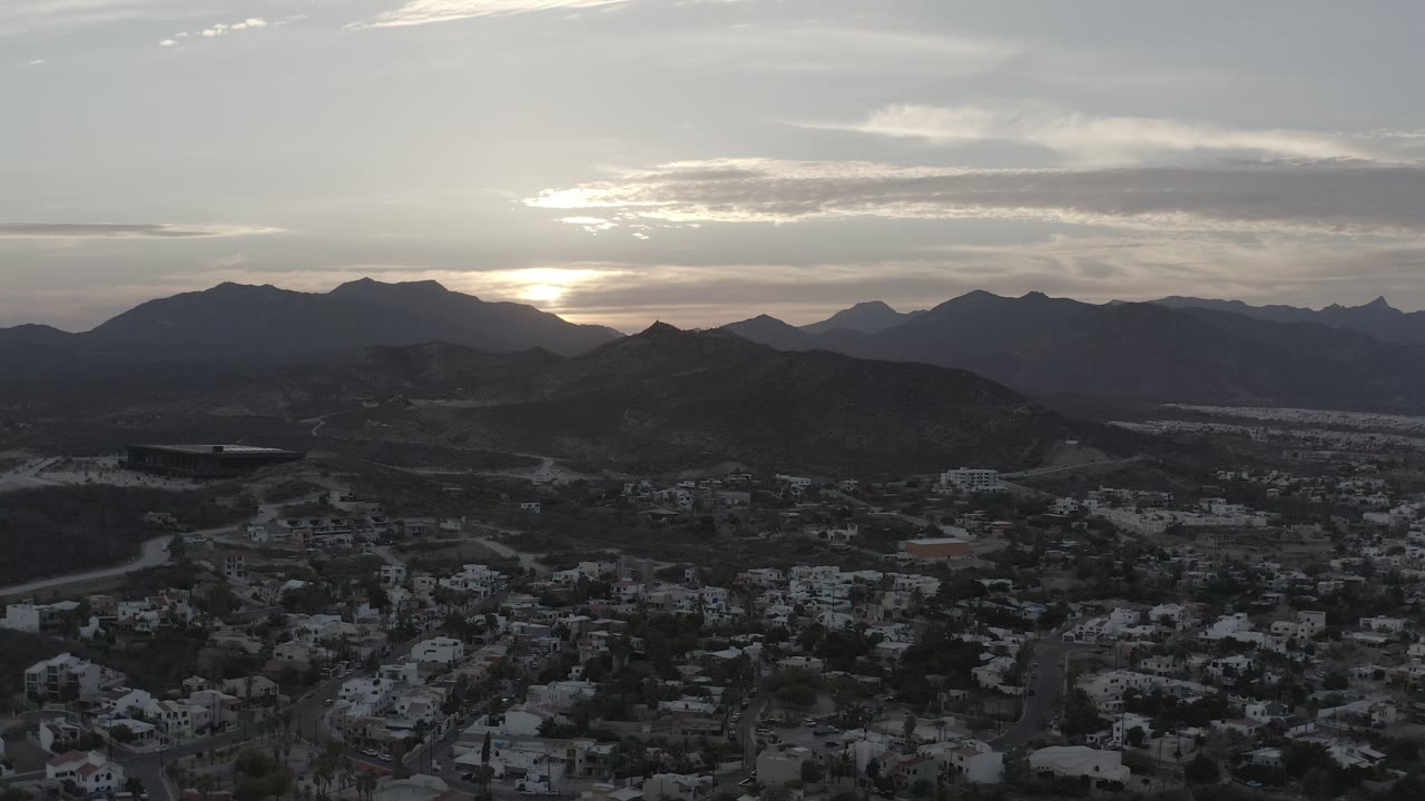 Los Cabos, Mexico sunset aerial with rooftops and roads in the lower frame, rounded hills in the middle distance, mountain silhouettes, clouds, and no visible people.
