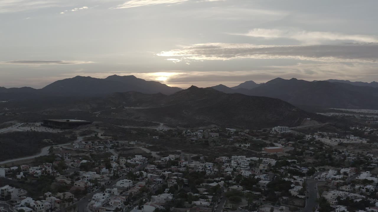 Los Cabos, Mexico appears from an elevated view with the low sun glowing behind the mountain ridge and residential streets visible in the darker foreground, with no visible people.