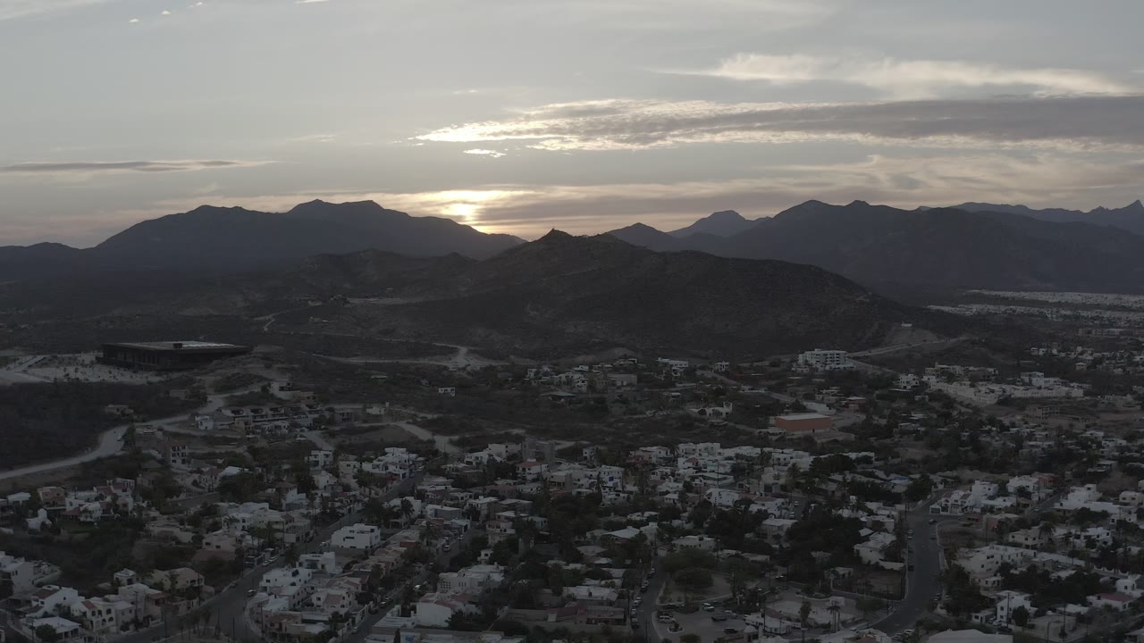 Elevated Los Cabos view with residential streets in shadow, rounded hills across the center, distant mountains along the horizon, and a pale sun glow filtered by clouds.
