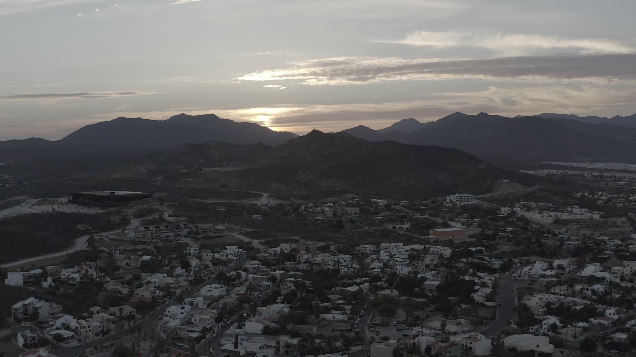 Los Cabos rooftops and roads fill the lower frame while dark hills and a cloud-covered sunset sit across the horizon, with no visible people.