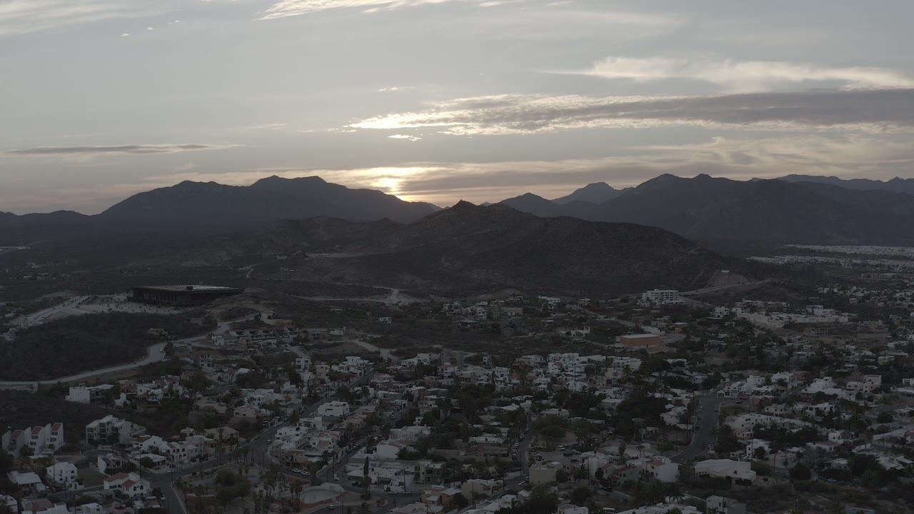 Wide aerial view of Los Cabos at dawn, with dark mountain ridges on the horizon, scattered buildings below, a bright clouded sky, and no visible people.