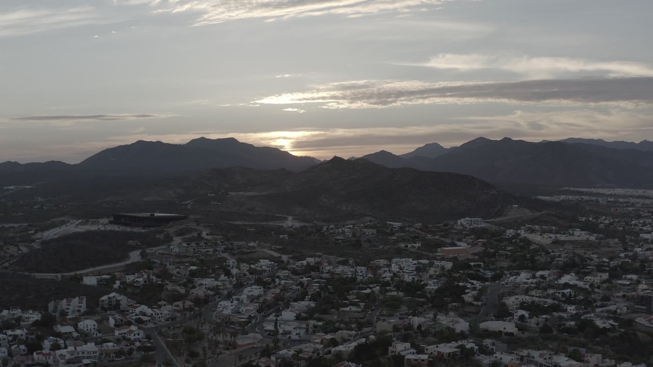 Wide elevated view in Los Cabos, Mexico, showing dark foothills, town buildings below, a bright morning horizon behind layered mountains, and no visible people.
