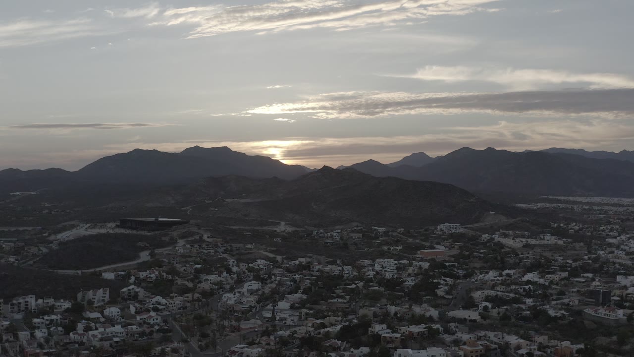 Los Cabos aerial frame with clustered light buildings in the foreground, dark hills across the middle, and the low sun partly hidden by clouds, with no visible people.
