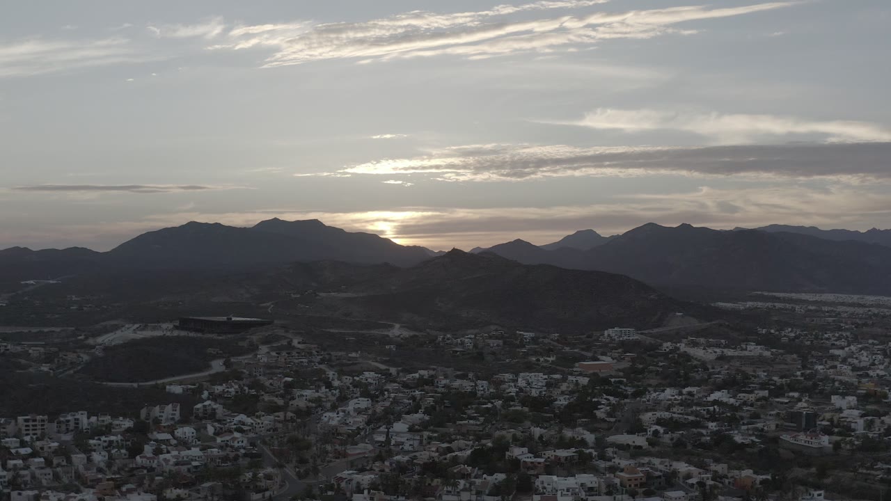 Los Cabos, Mexico appears from an elevated angle with city blocks in shadow, rounded hills in the midground, a mountain horizon, and no visible people.