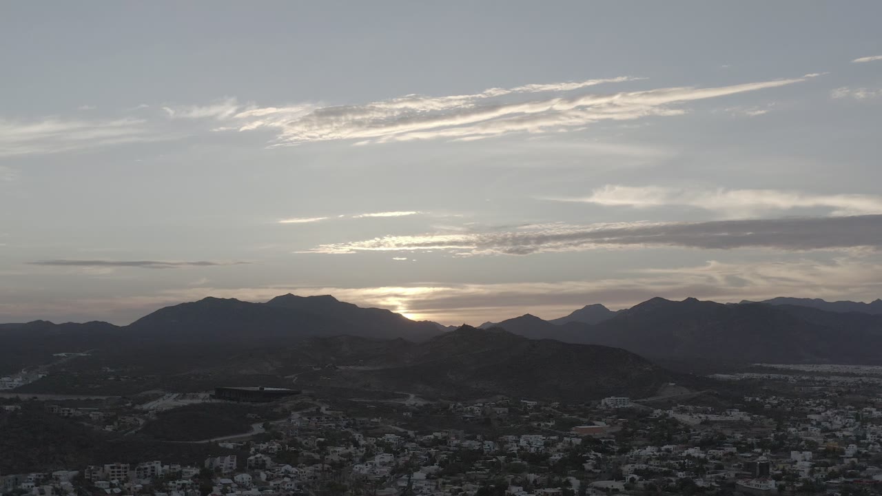 Opening frame of Los Cabos, Mexico with a dark town below, layered mountain silhouettes, streaked clouds, and no visible people.