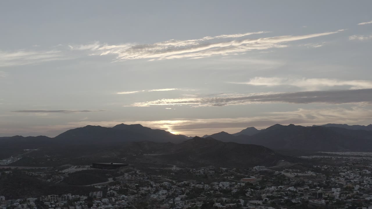 Los Cabos, Mexico skyline view with the sun tucked behind the mountain ridge, a dark urban foreground, thin bright cloud streaks, and no visible people.
