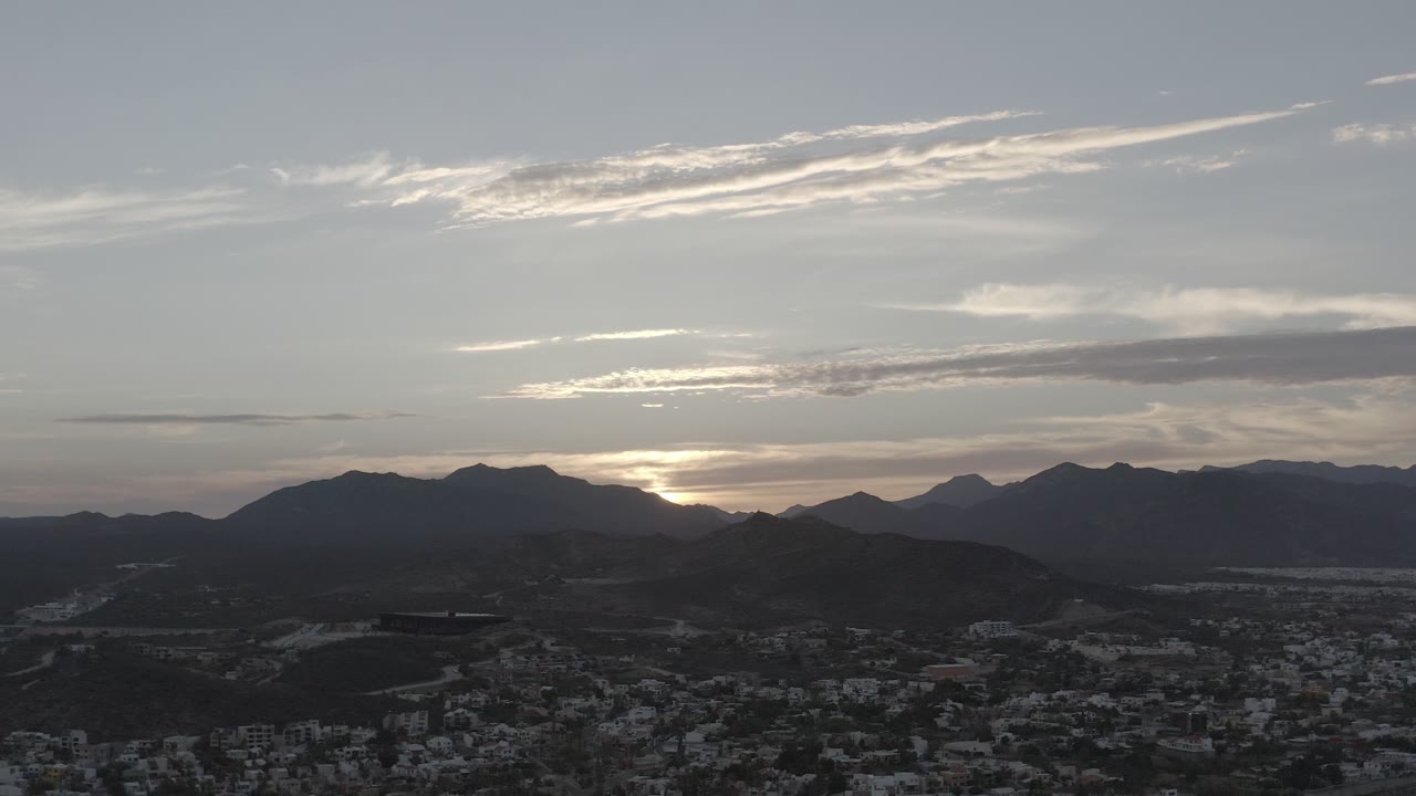 Los Cabos mountain skyline with a bright band of daylight behind the ridges, scattered city blocks below, and no visible people.