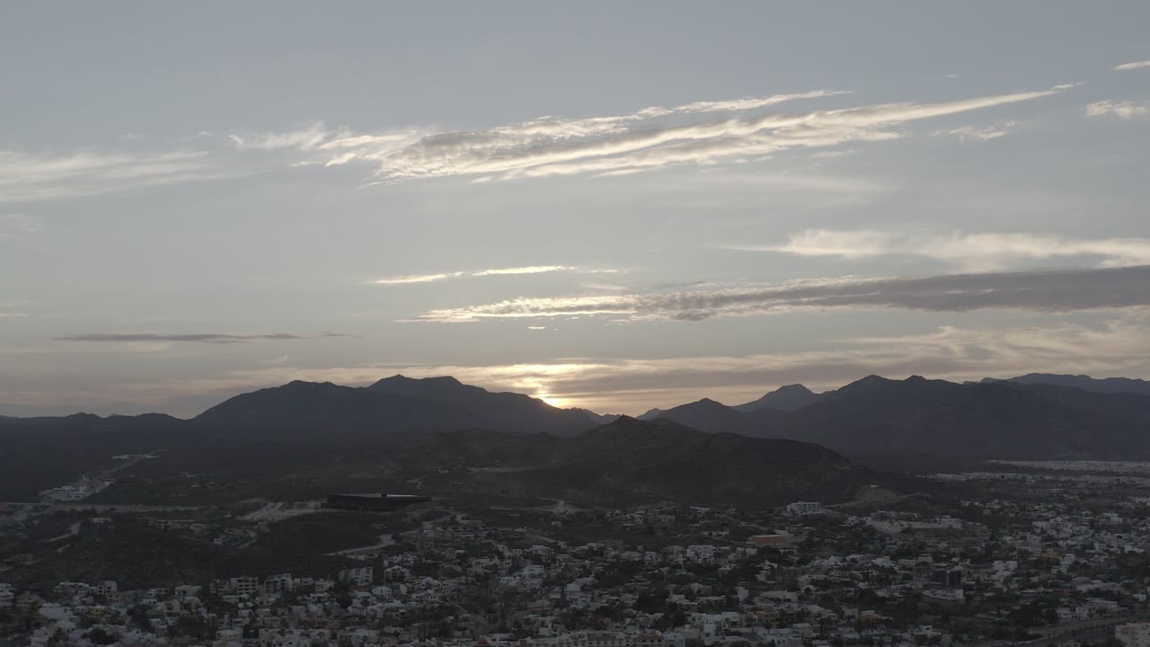 Opening frame of Los Cabos, Mexico, with the sun low behind dark mountains, pale clouds across the sky, a shadowed town below, and no visible people.