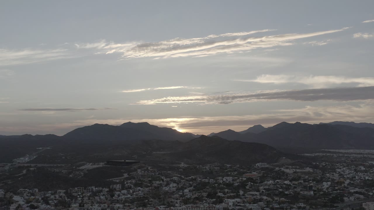 Los Cabos sunset view one second in, showing a bright band of light behind the ridge, layered mountain silhouettes, cloud streaks, and no visible people.