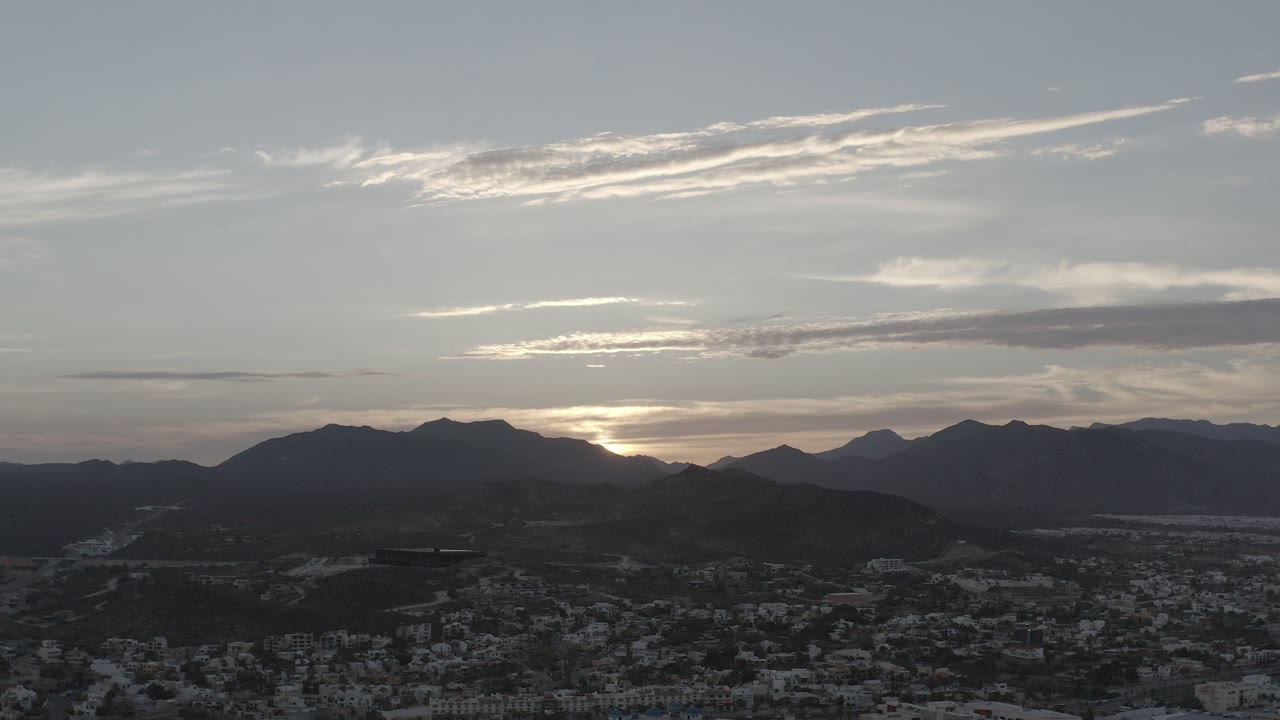 Wide aerial view in Los Cabos, Mexico, with dark mountain ridges, low sun behind the horizon, scattered clouds, town buildings below, and no visible people.