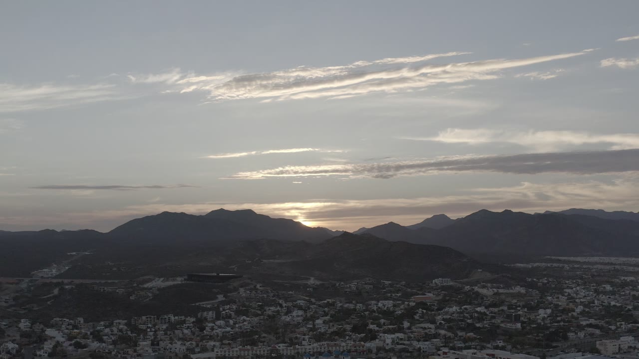 Aerial Los Cabos view with the sun pressed lower behind the mountain line, gray clouds across the sky, shadowed hills, town blocks at the bottom, and no visible people.