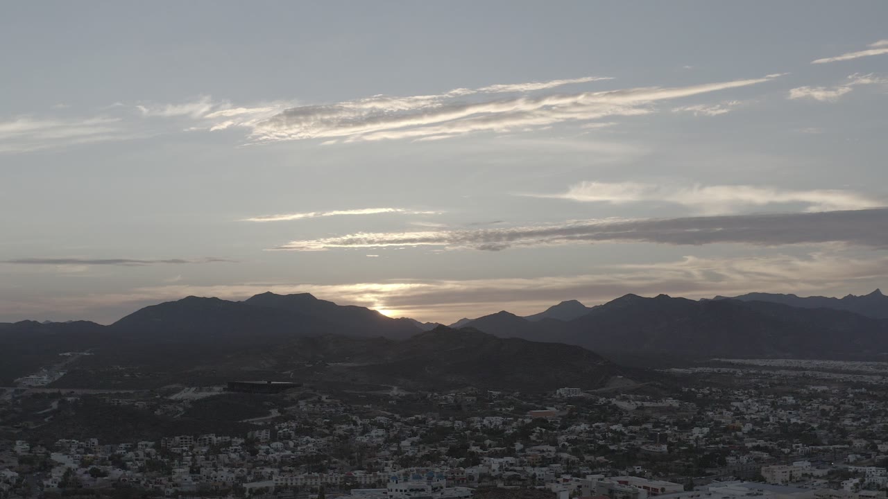 Los Cabos sunset view with dark mountain ridges, bright cloud bands, a town spread across the lower frame, and no visible people.