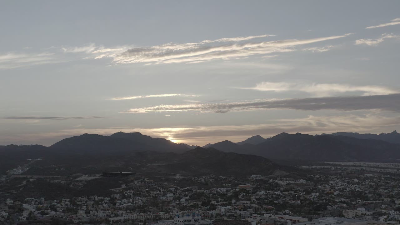 Los Cabos overlook frame with the sun tucked behind the mountain ridge, pale clouds across the sky, city blocks below, and no visible people.