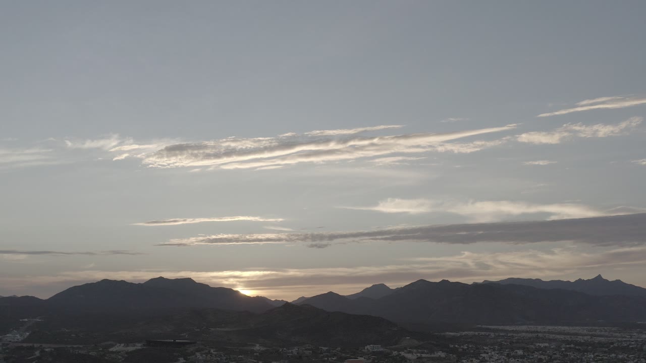 Opening frame of a Los Cabos, Mexico sunset with the sun partly hidden by a mountain ridge, pale cloud bands above, and no visible people.