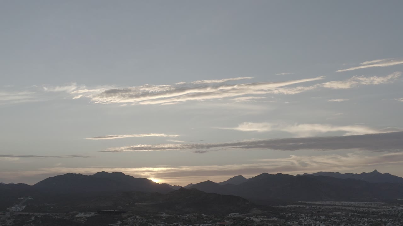 Opening frame of a Los Cabos, Mexico sunset, with the sun low behind dark mountain ridges, layered clouds above, a distant city below, and no visible people.