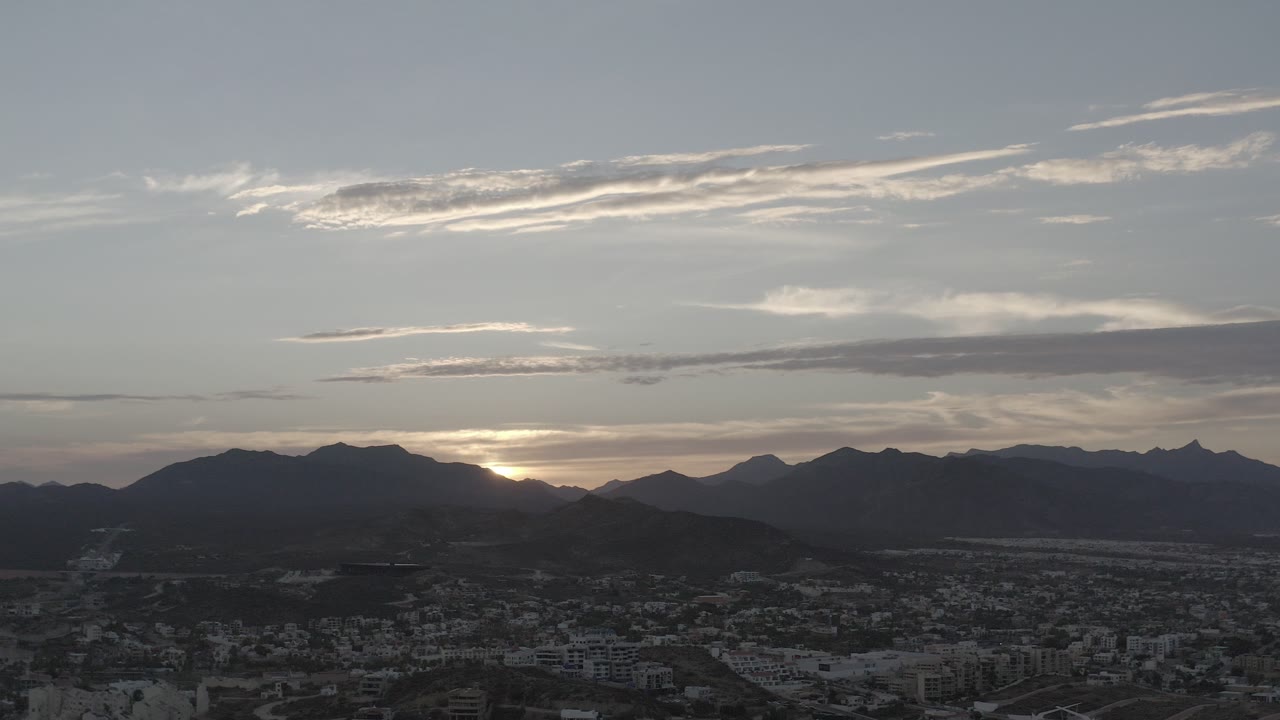 Wide elevated view of Los Cabos, Mexico, with a town in shadow below dark mountain ridges, pale sunset light behind the peaks, streaked clouds overhead, and no visible people.