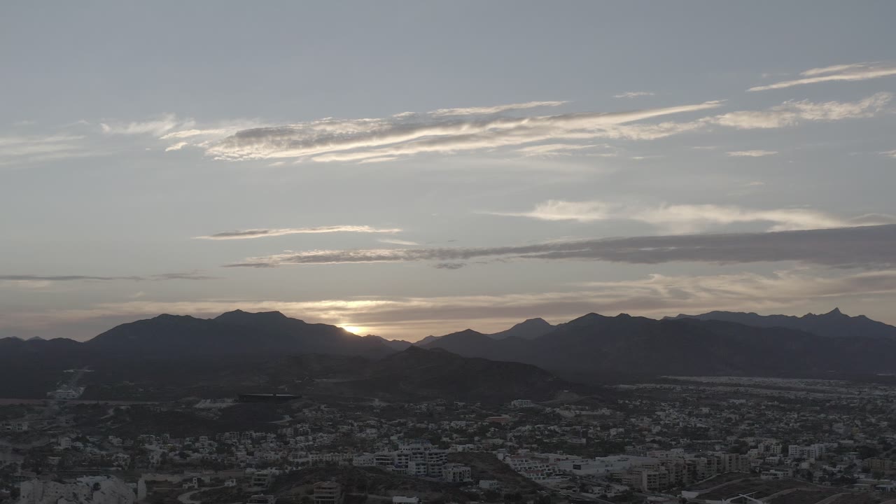 Elevated Los Cabos, Mexico landscape with dense town blocks in the foreground, a dark mountain skyline across the middle, thin bright clouds above, and no visible people.