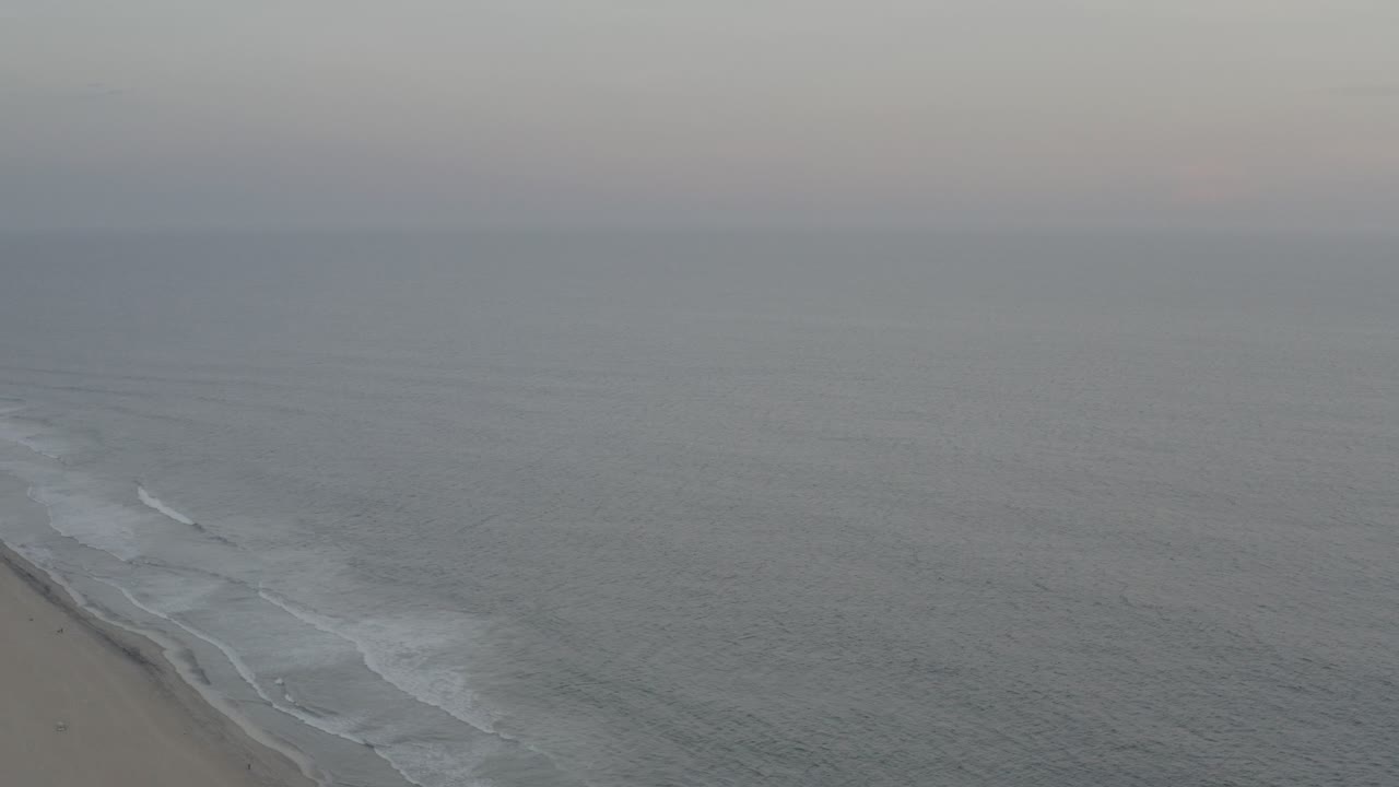 Aerial view in Los Cabos, Mexico, with gray ocean water, pale surf breaking along a sandy beach, a hazy horizon, and no visible people.