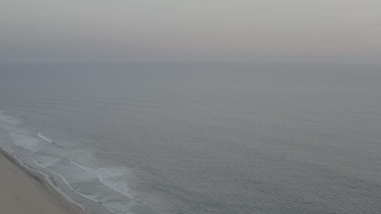 Wide Los Cabos aerial frame where rippled gray-blue water dominates, with a thin diagonal strip of surf and sand at the lower left and no visible people.