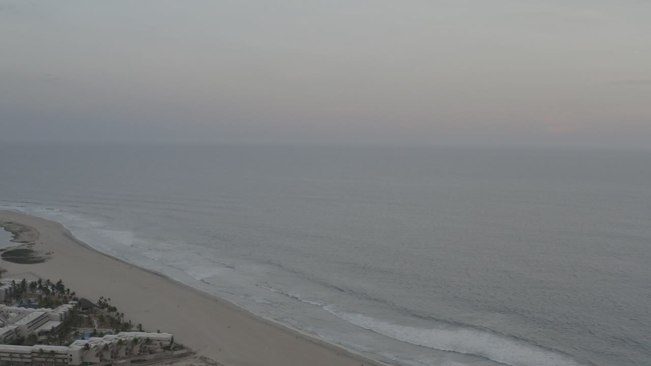 Opening aerial view in Los Cabos, Mexico, with a wide sandy shore, gray-blue ocean, white surf bands, pale sky, and no visible people.