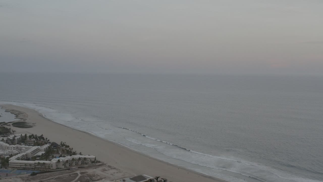 Aerial view of beach, surf, ocean horizon, and beachfront buildings.