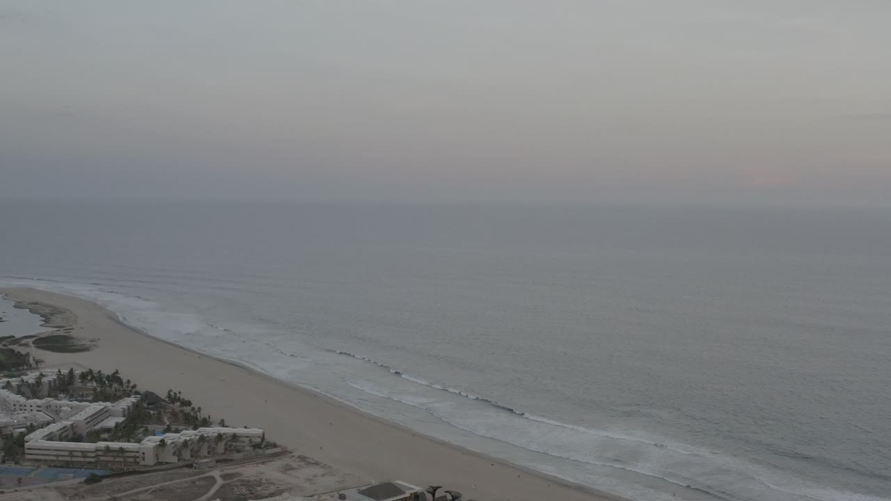 A Los Cabos shoreline frame shows the sandy beach curving along the left side, thin white wave lines offshore, low buildings near the bottom edge, and no visible people.