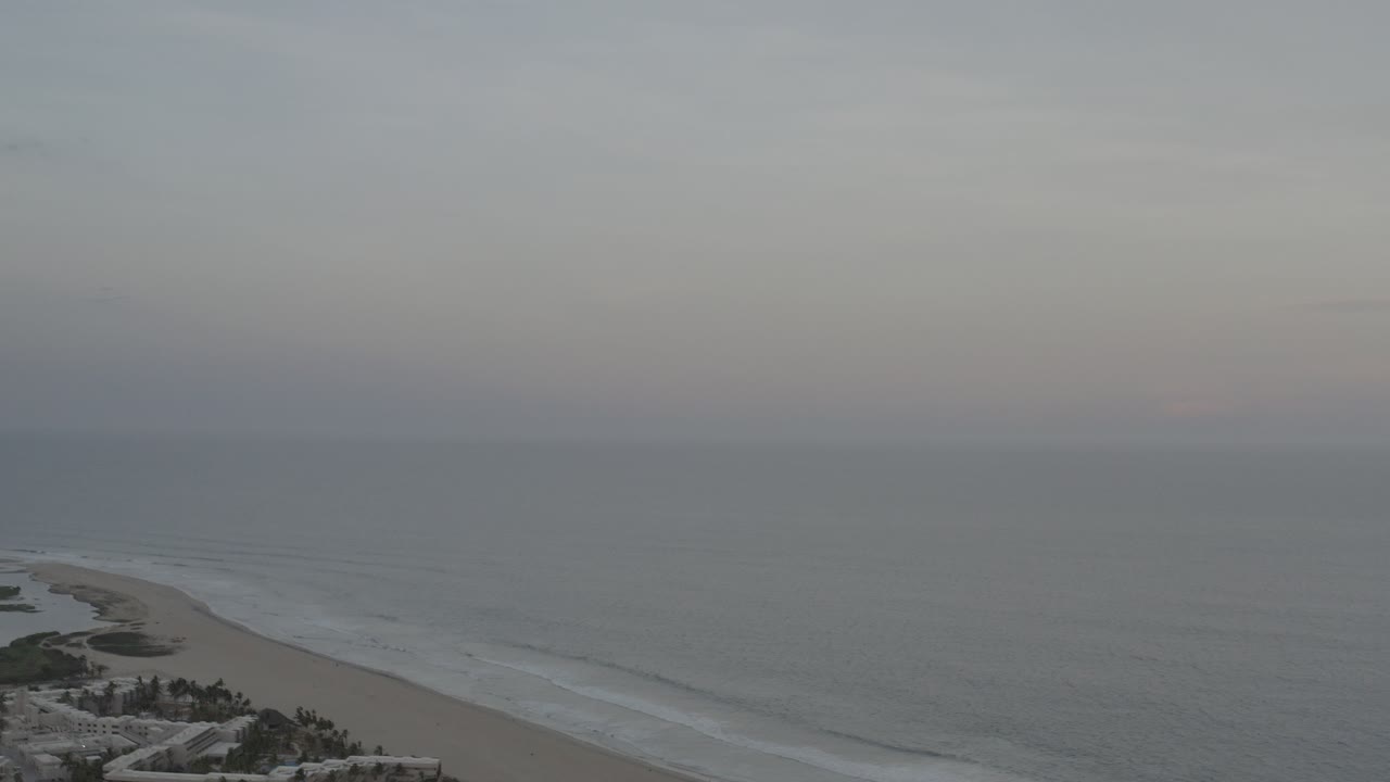 Opening aerial view of the Los Cabos, Mexico coast with gray ocean, pale beach, distant surf lines, low clouds, and no visible people.