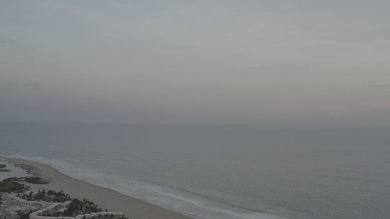Aerial frame of Los Cabos, Mexico showing a curved sandy shoreline, white wave bands, gray water, cloudy sky, and no visible people.