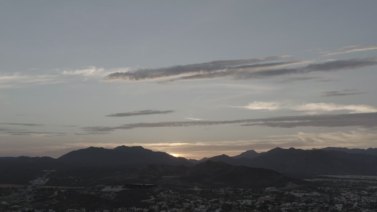 Wide elevated view in Los Cabos, Mexico, with the sun tucked behind a dark mountain ridge, blue-gray clouds overhead, a shadowed town below, and no visible people.