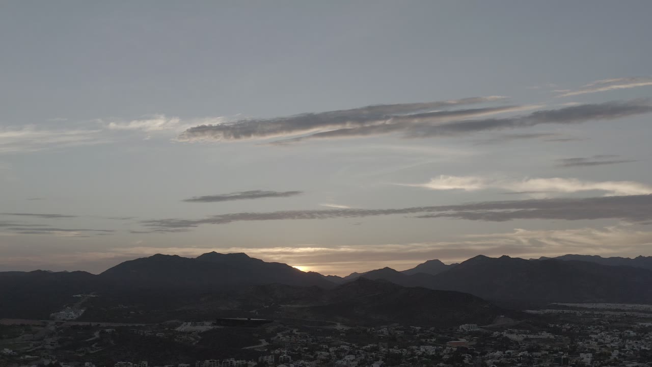 Los Cabos aerial frame with layered dusk clouds above a dark ridge line and a dense residential area fading into shadow, with no visible people.