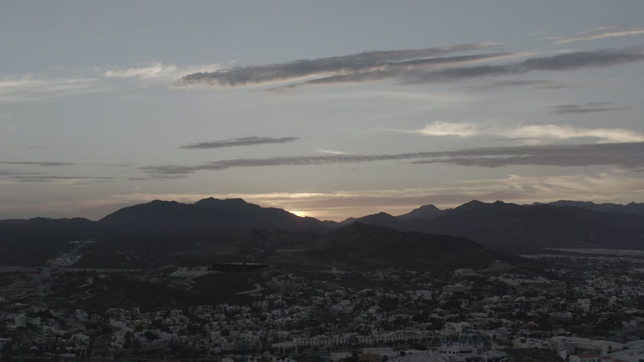 Los Cabos mountain ridges sit under layered gray clouds, with a compact settlement visible in the lower frame and no visible people.