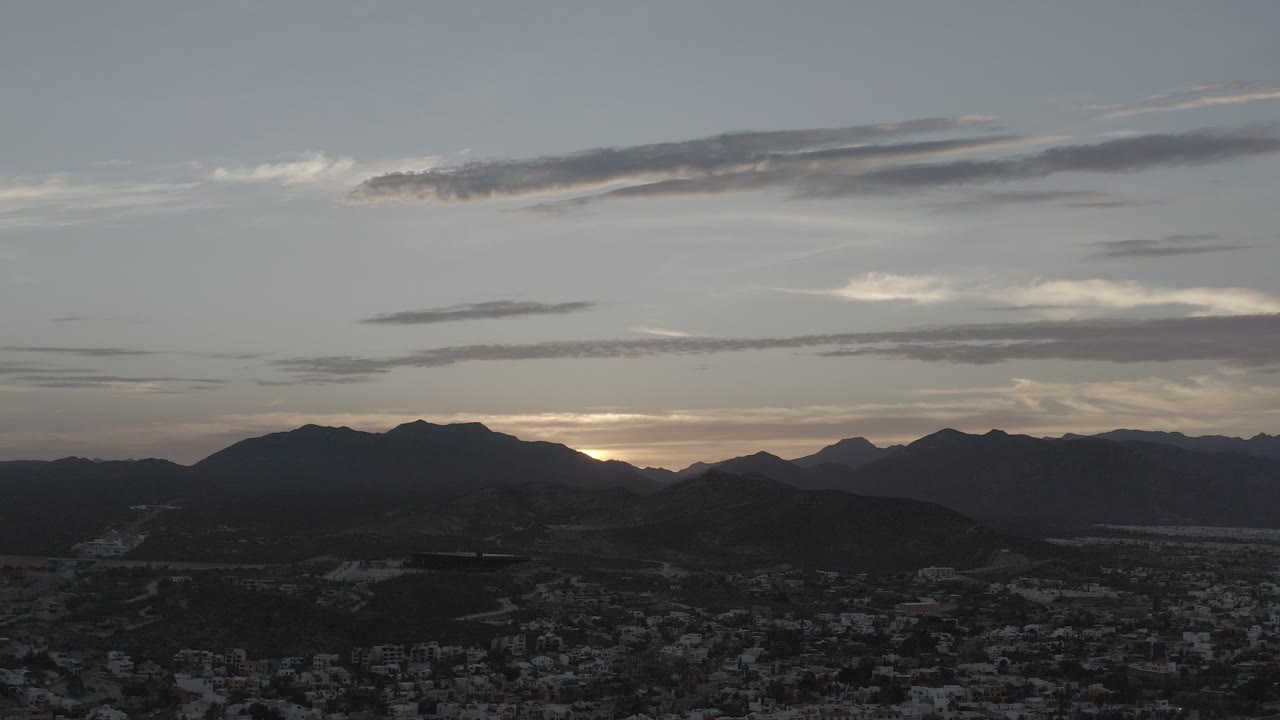 Los Cabos, Mexico dusk view with dark mountain silhouettes, a dim city foreground, layered clouds, and no visible people.