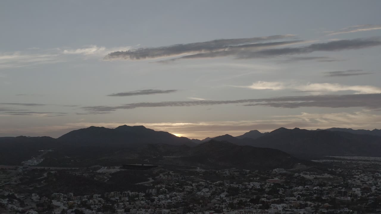 Los Cabos, Mexico appears from an elevated viewpoint, with a shadowed city below a broad mountain ridge and pale clouds, no visible people.