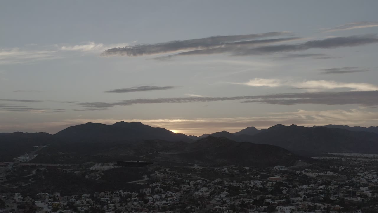 Early frame over Los Cabos, Mexico showing the silhouetted mountains slightly brighter at the horizon, gray cloud bands across the sky, dark foreground hills, town buildings below, and no visible people.