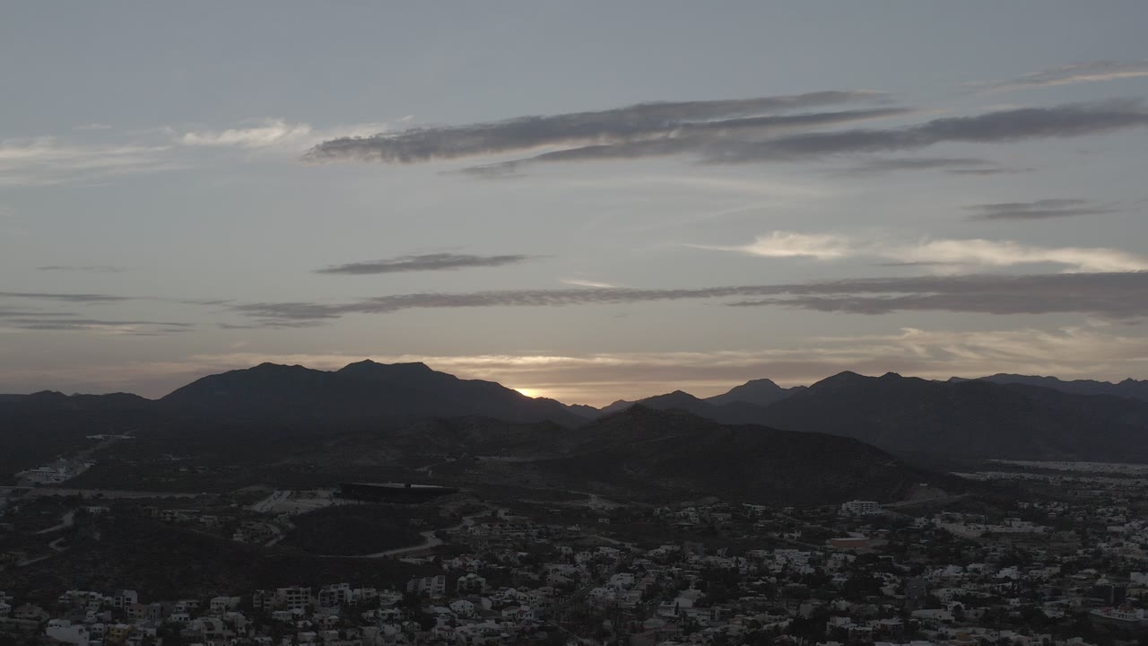 Opening frame of a Los Cabos, Mexico sunset with dark mountain silhouettes, a faint town below, layered clouds above, and no visible people.