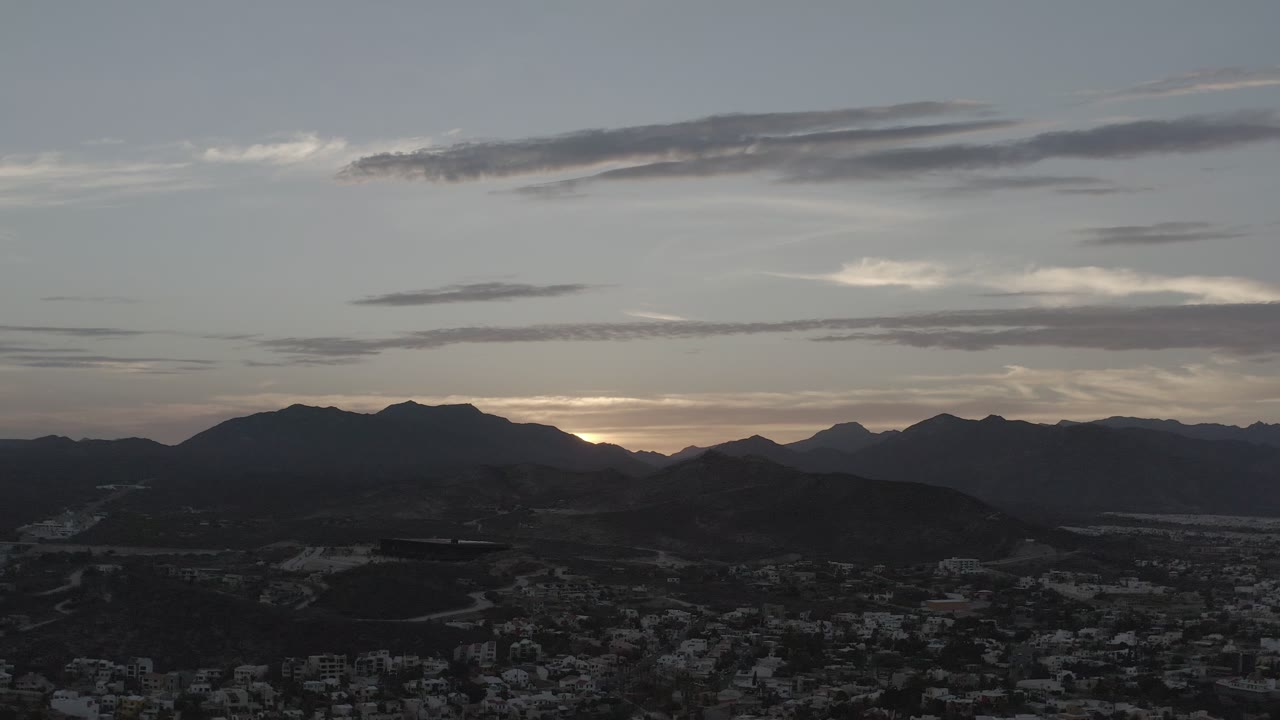 A Los Cabos, Mexico dusk frame with a pale sky, elongated clouds, dark hills across the center, dim buildings at the bottom, and no visible people.