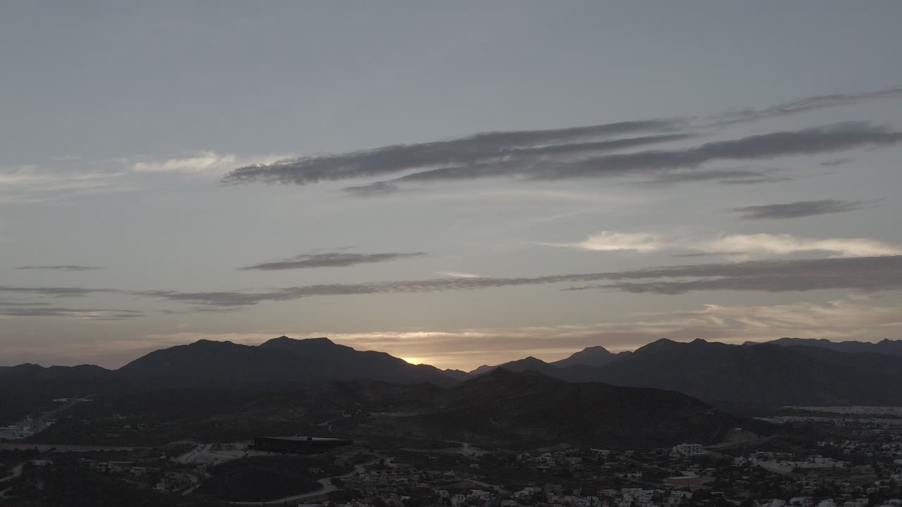 Los Cabos, Mexico dusk view with dark hill and mountain silhouettes under gray-blue cloud bands, warm horizon light, and no visible people.