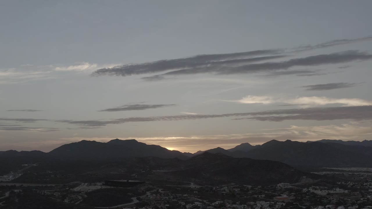 Los Cabos, Mexico evening sky with long dark clouds, a pale sunset strip behind the mountains, shadowed hills below, and no visible people.