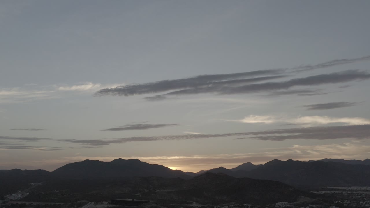 Wide dusk view in Los Cabos, Mexico with dark mountain ridges along the bottom, layered gray clouds overhead, warm light at the horizon, and no visible people.